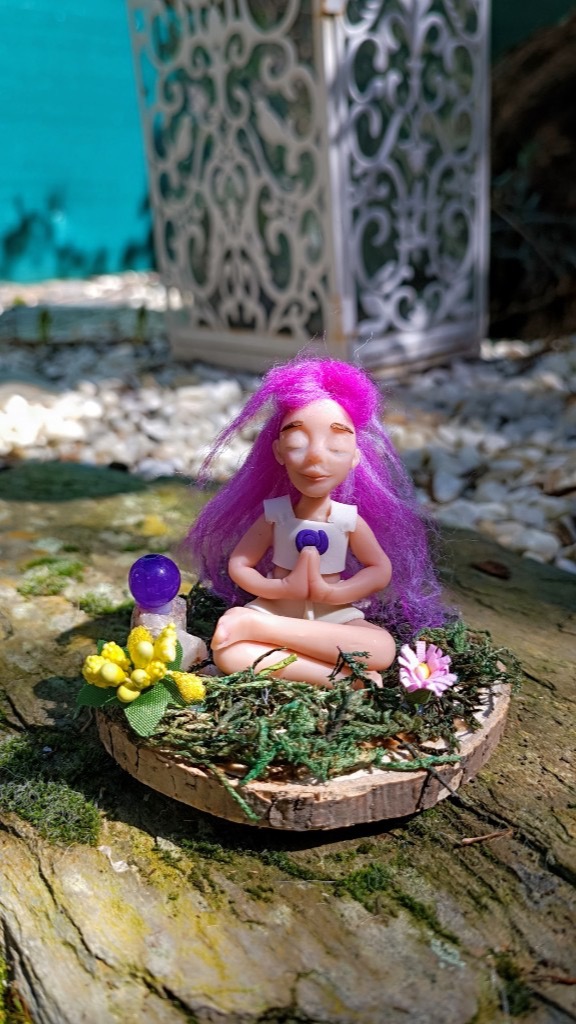 Figura meditando con pelo morado sobre rodaja de madera con musgo y flores