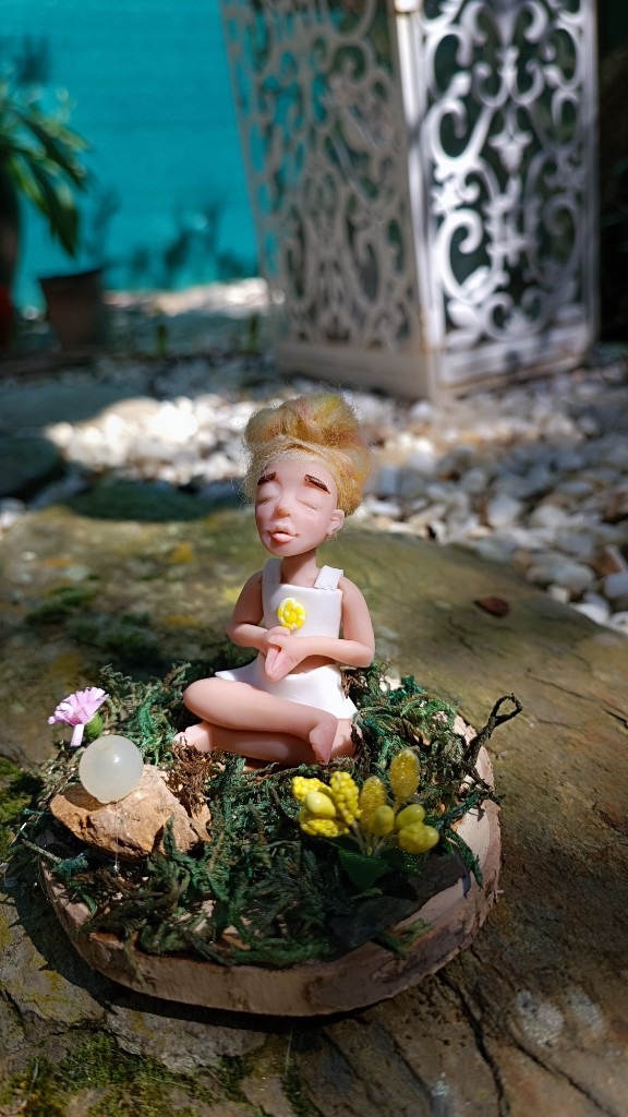 Figura meditando con moño rubio, vestido blanco y flores en rodaja de madera