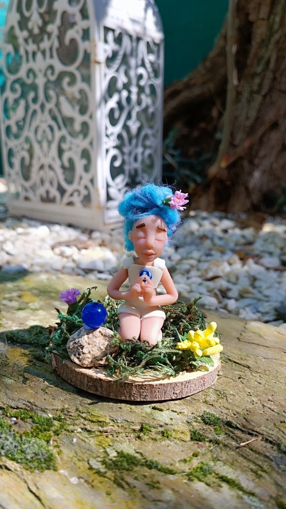 Figura meditando con pelo azul rizado, flor rosa y canica azul en la base