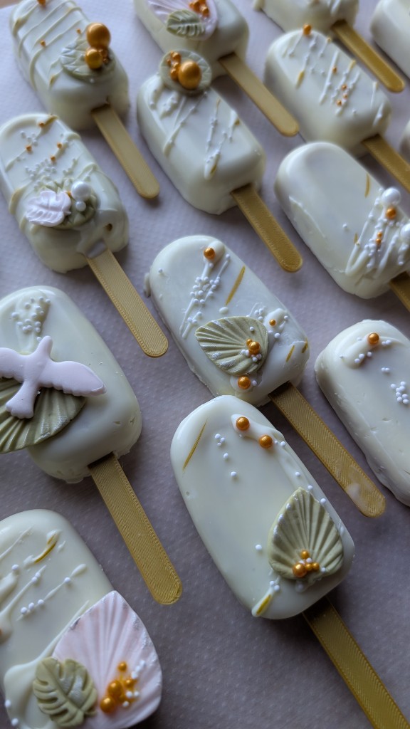 Cakesicles blancos decorados con hojas, perlas doradas y paloma para comunión o boda